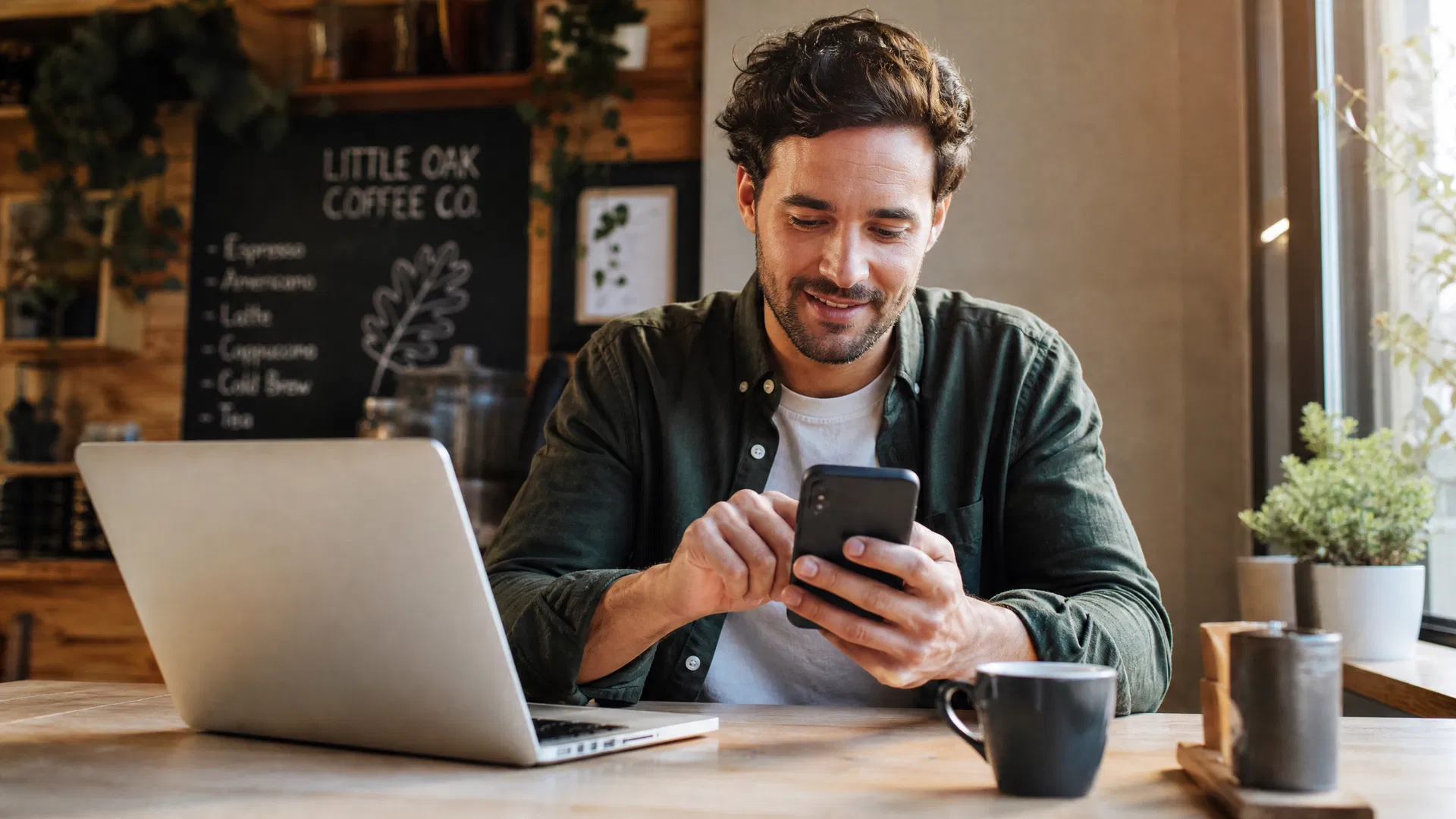 Small business owner updating their Google Business Profile services section on a smartphone at a cafe with a laptop nearby